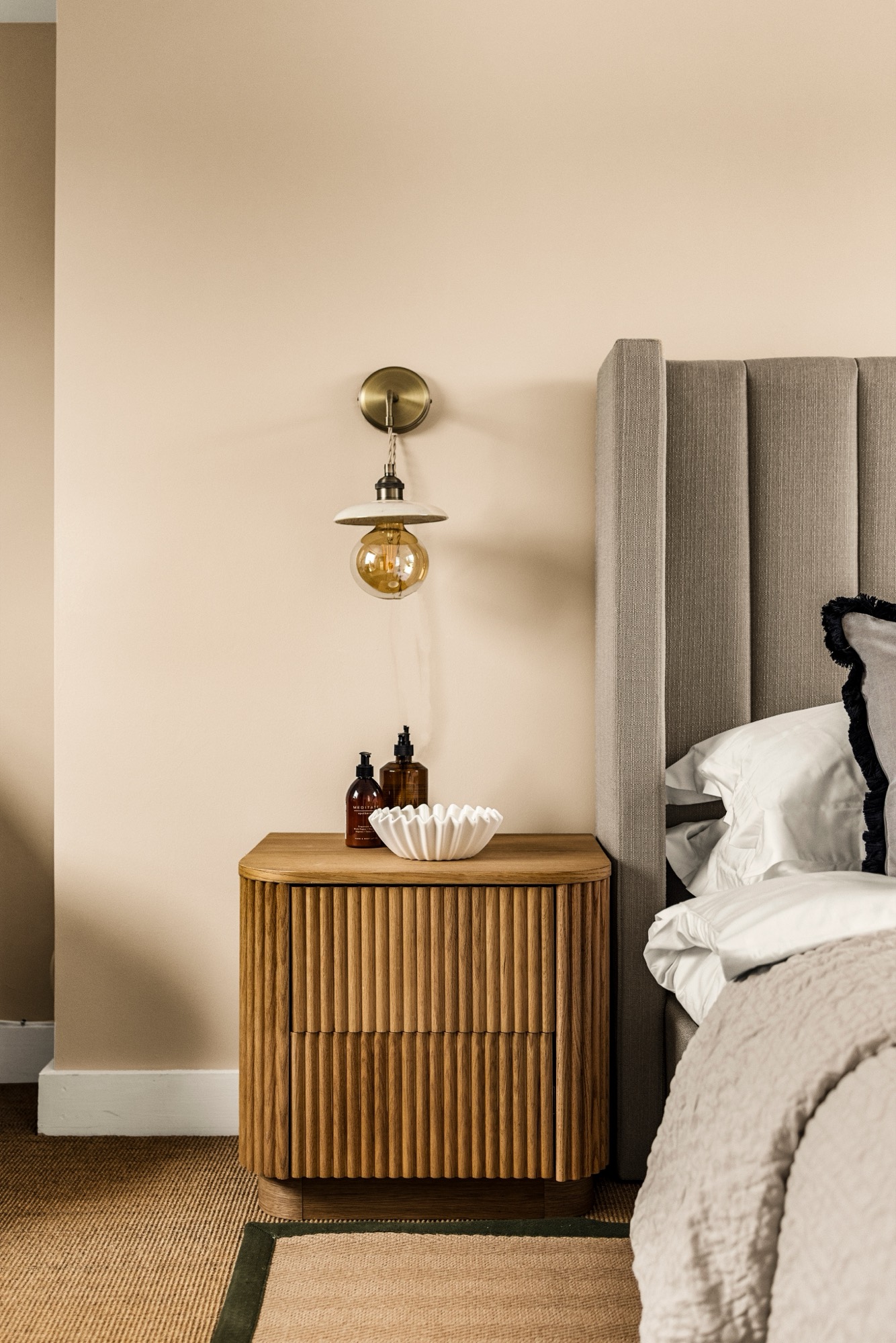 Bedroom detail photography: ribbed oak bedside table, brass wall light and upholstered headboard