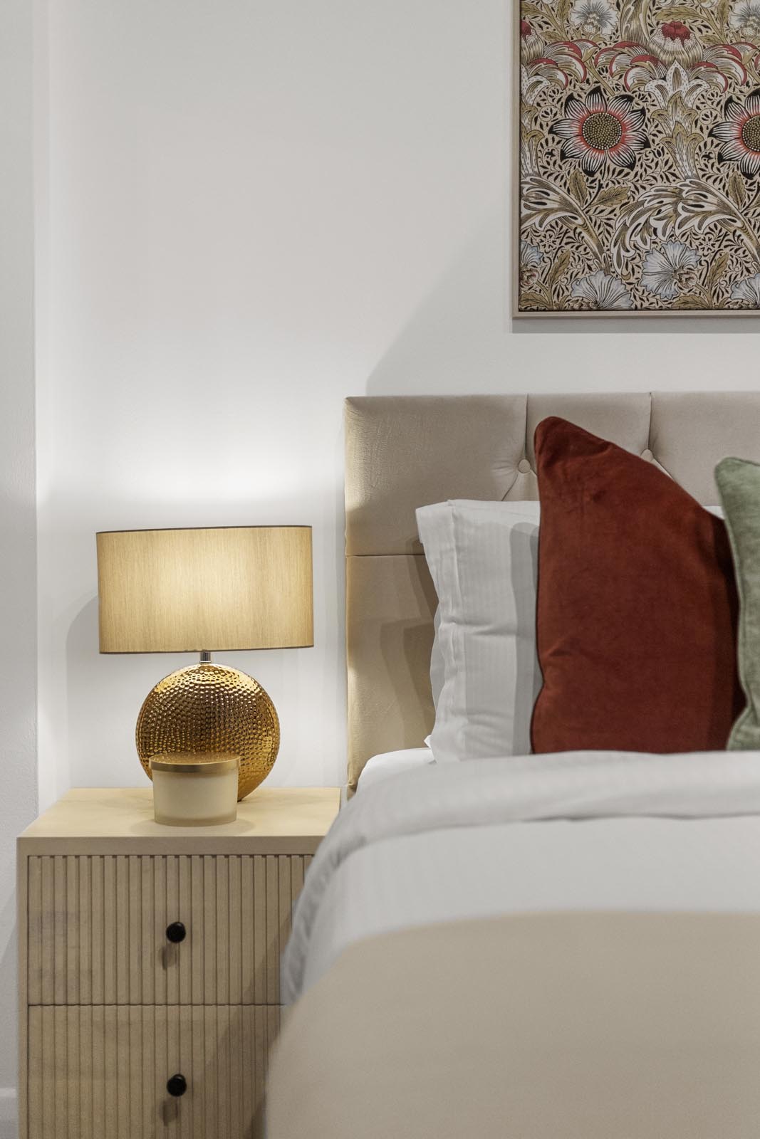 Bedroom detail photography in a Kensington flat: brass lamp on a ribbed bedside table, tapestry art and rust cushion